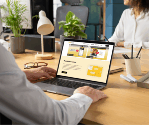 Persoon die een laptop gebruikt op een bureau in een moderne kantooromgeving, met digitale content en LifeCheck-website zichtbaar op het scherm, planten, lamp en schrijfwaren op de achtergrond.
